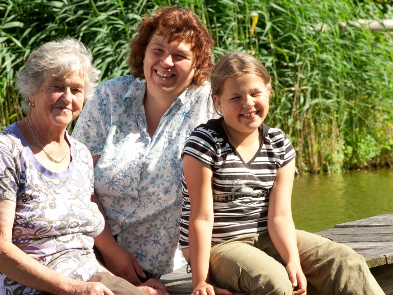 Drei Frauen aus drei Generationen - von alt bis jung - sitzen gemeinsam auf einem Steg an einem See