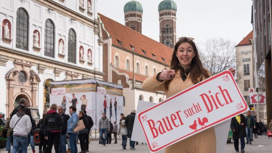 Eine junge Teilnehmerin des Herrschinger Grundkurses mit dem Schild Bauer sucht Dich in der Hand vor der Frauenkirche in München