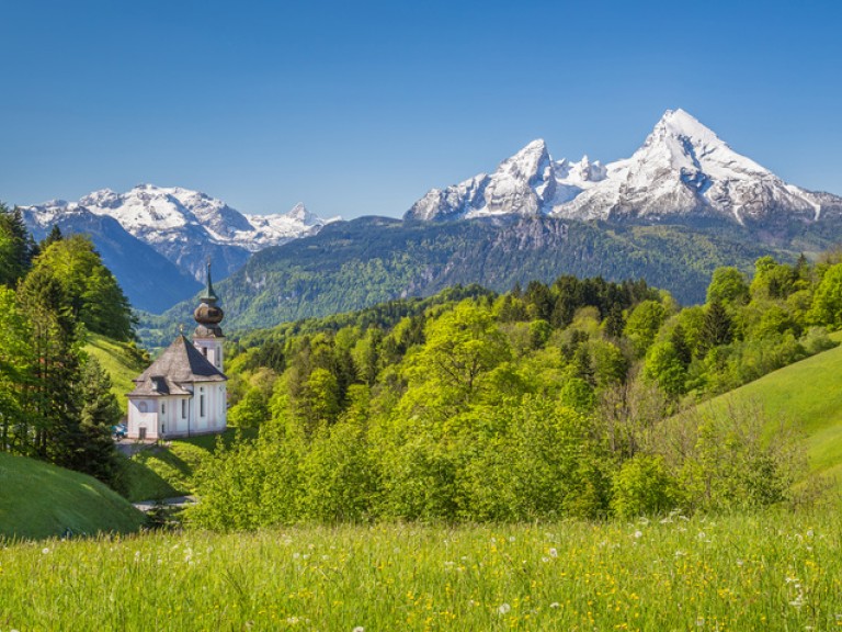 Voralpenlandschaft in Bayern mit Wäldern und Kirche, im Hintergrund schneebedeckte Berge