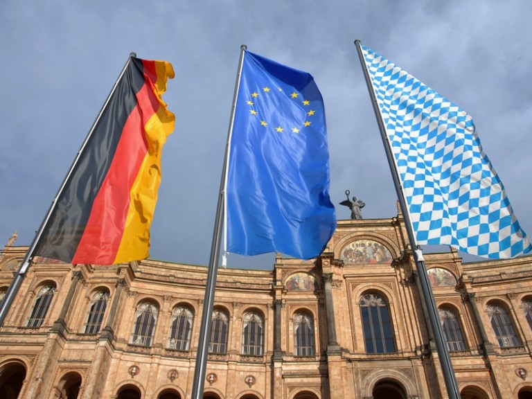 Die Deutschland-, Eu-, und Bayernfahne vor einem historischen Gebäude