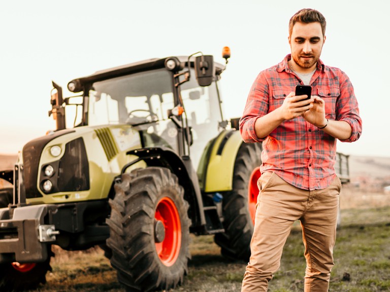 Ein junger Landwirt steht mit seinem Smartphone neben seinem Traktor
