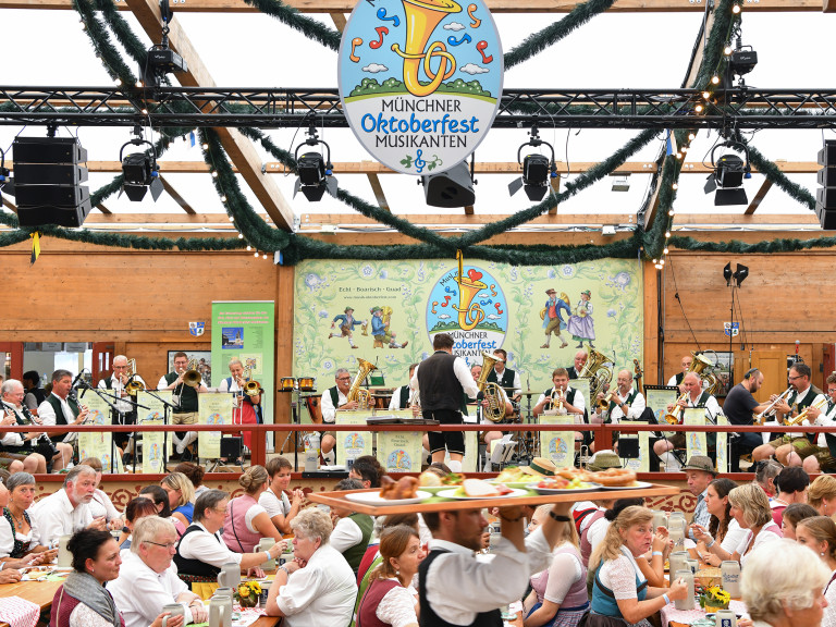 Blick ins voll besetzte Festzelt. Im Hintergrund spielen auf der Bühne die Münchner Oktoberfest Musikanten. Im vordergrund trägt ein Ober ein volles Tablett vorbei.
