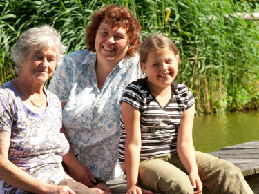 Drei Frauen aus drei Generationen - von alt bis jung - sitzen gemeinsam auf einem Steg an einem See