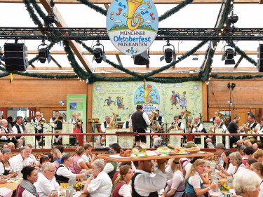 Blick ins voll besetzte Festzelt. Im Hintergrund spielen auf der Bühne die Münchner Oktoberfest Musikanten. Im vordergrund trägt ein Ober ein volles Tablett vorbei.