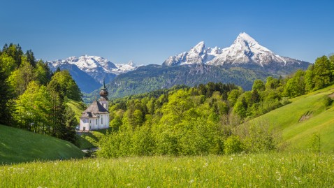 Voralpenlandschaft in Bayern mit Wäldern und Kirche, im Hintergrund schneebedeckte Berge