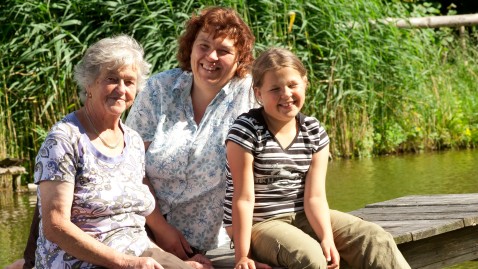 Drei Frauen aus drei Generationen - von alt bis jung - sitzen gemeinsam auf einem Steg an einem See