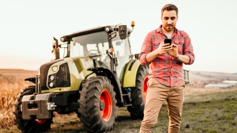 Ein junger Landwirt steht mit seinem Smartphone neben seinem Traktor 
