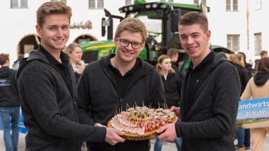 Drei junge Teilnehmer des Herrschinger Grundkurses halten einen Holzteller mit Kostproben an Wurst und Speck