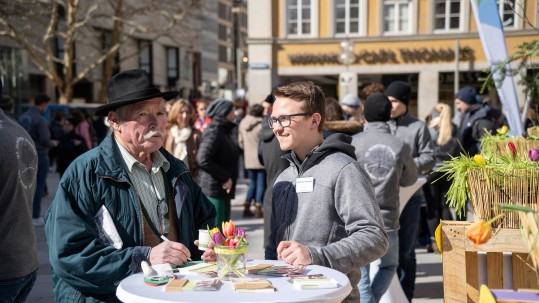 Münchner mit beeindruckendem Schnauzbart spricht mit jungem Bauern auf dem Marienplatz