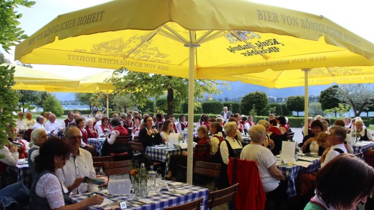 Chorsängerinnen und Sänger in ihren Trachten sitzen bei strahlendem Sonnenschein im Biergarten des Festspielhauses