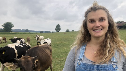 Eine Frau schaut in die Kamera, hinter ihr stehen ein paar Kühe auf einer Weide