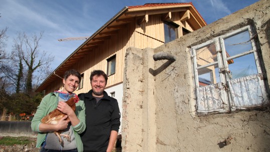 Frau mit Huhn und Mann stehen vor einem Haus mit Baustelle